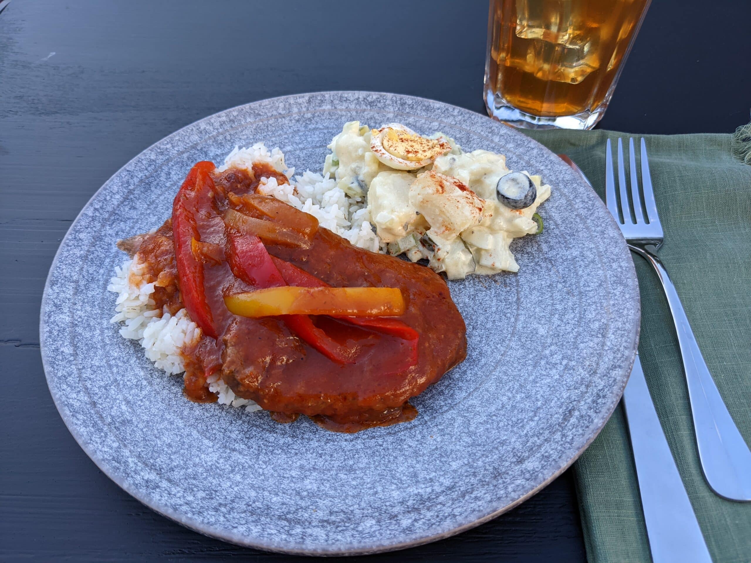 Country style cube steak on a grey plate with potato salad, in the background is a glass of iced tea, a knife and fork