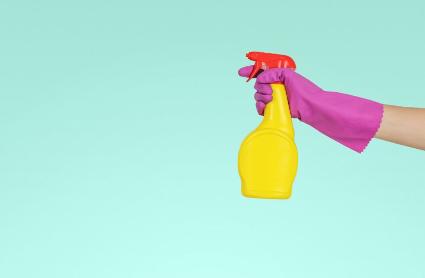 Blue background, hand in pink cleaning glove holding yellow and red spray bottle