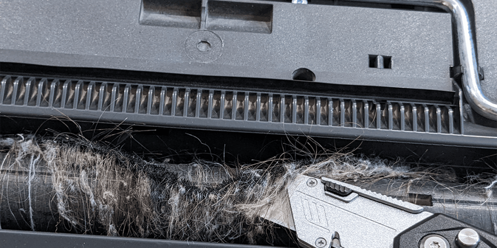 Long hair wrapped around the brush bar of a vacuum. A box cutter is slicing through the hair